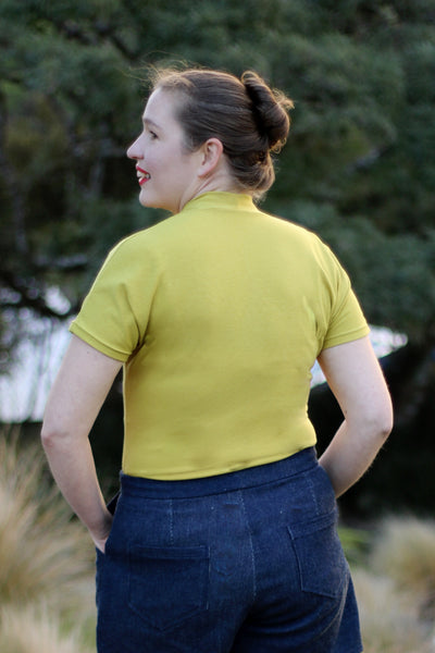 Leimomi, a white woman with her hair in a bun, is shown wearing the Scroop Miramar Top with short sleeves in yellow cotton lycra jersey.  She shows the back view of the top