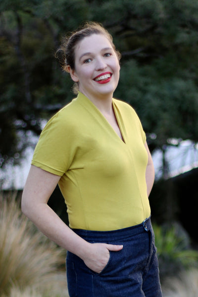 Leimomi, a white woman with her hair in a bun, is shown wearing the Scroop Miramar Top with short sleeves in yellow cotton lycra jersey