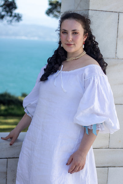 Woman in a white 17th c shift with puffed sleeves standing by a stone wall with ocean view