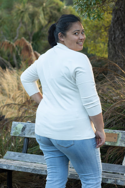 Rose, a Pasifika woman with her hair in a ponytail, is shown wearing the Scroop Miramar Top with 3/4 length sleeves in ivory ponte knit.  She faces away from the camera and looks over her shoulder.