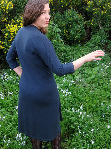 Leimomi, a white woman with her hair in a braid, is shown wearing the Scroop Miramar Dress with 3/4 length sleeves in slate blue wool knit.  She stands with her back to the camera and looks over her shoulder.